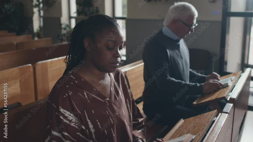 High angle zoom in of modern young African American woman sitting on bench in Catholic church listening to pastor and reading the Bible High angle zoom in of modern young African American woman sitting on bench in Catholic church listening to pastor and reading the Bible