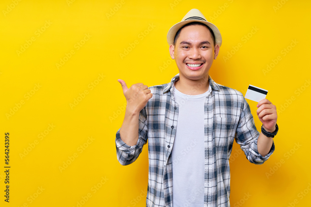 Cheerful Asian ethnicity young man 20s wearing casual clothes hat and holding in hand credit bank card, pointing his thumb aside isolated on yellow background studio portrait. People lifestyle concept Cheerful Asian ethnicity young man 20s wearing casual clothes hat and holding in hand credit bank card, pointing his thumb aside isolated on yellow background studio portrait. People lifestyle concept