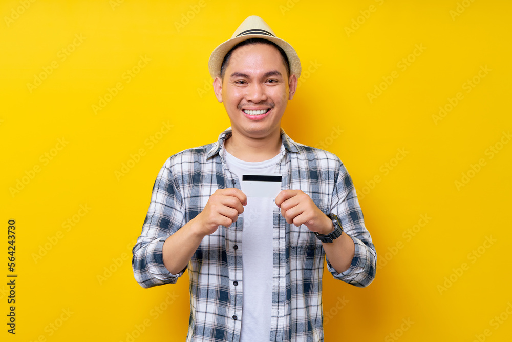 Smiling satisfied Asian ethnicity young man 20s wearing casual clothes hat holding credit bank card, looking confident at camera isolated on yellow background studio portrait. People lifestyle concept
