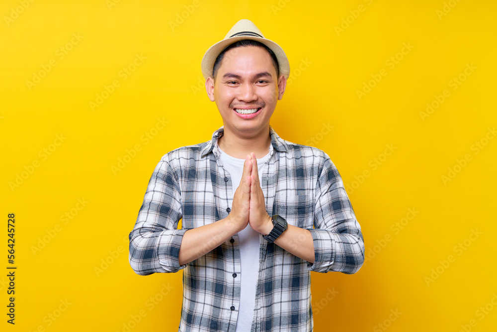 smiling Young handsome ethnic Asian man 20s wearing casual clothes hat showing namaste gesture while looking confident at the camera isolated on yellow background. People lifestyle concept