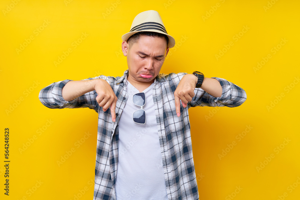Sadness Young handsome ethnic Asian man 20s wearing casual clothes hat standing pointing index fingers down on mock up copy space isolated on yellow background. People lifestyle concept Sadness Young handsome ethnic Asian man 20s wearing casual clothes hat standing pointing index fingers down on mock up copy space isolated on yellow background. People lifestyle concept
