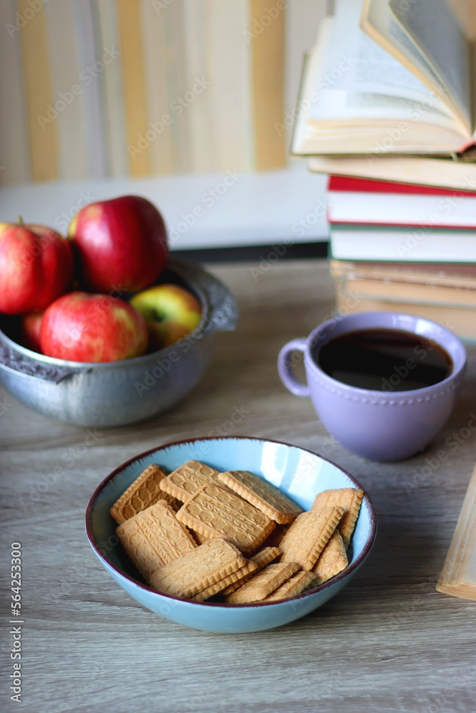 Stack of books, e-reader, reading glasses, bowl of biscuits, apples and cup of tea on the table. Bookshelf in the background. Selective focus.