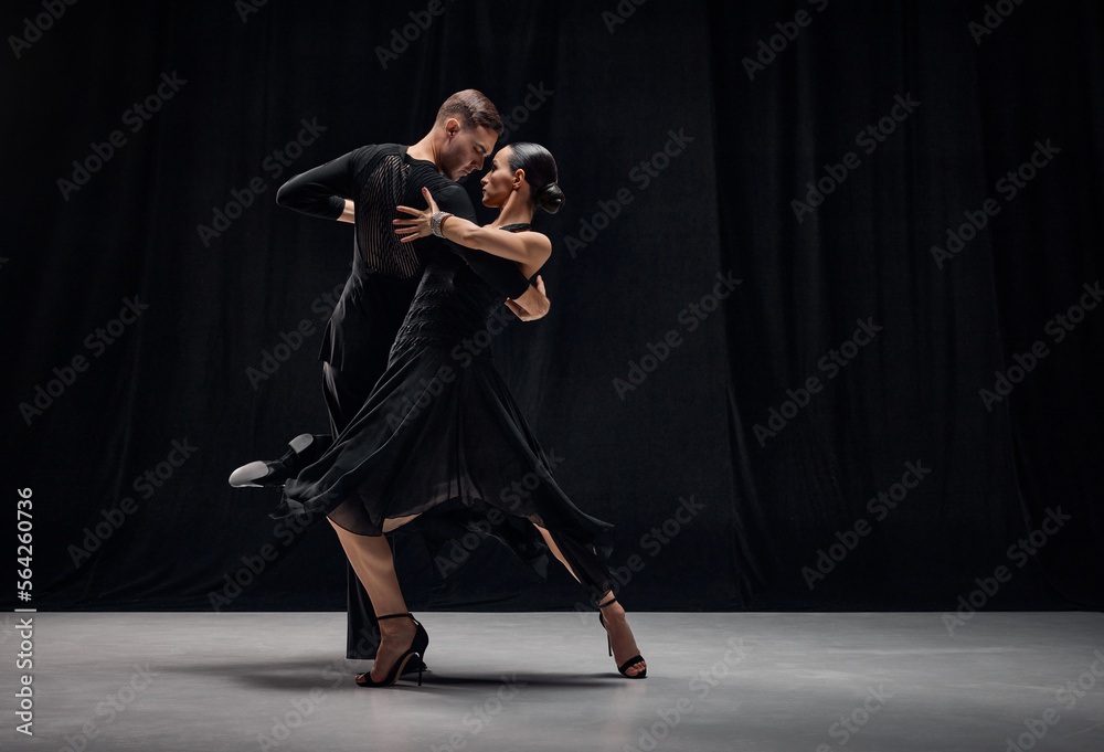 Man and woman, professional tango dancers performing in black stage costumes over black background. Expression of togetherness. Concept of hobby, lifestyle, action, motion, art, dance aesthetics