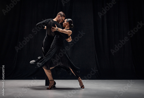 Man and woman, professional tango dancers performing in black stage costumes over black background. Expression of togetherness. Concept of hobby, lifestyle, action, motion, art, dance aesthetics