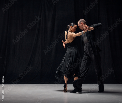Man and woman, professional tango dancers performing in black stage costumes over black background. Elegance, romance and passion. Concept of hobby, lifestyle, action, motion, art, dance aesthetics