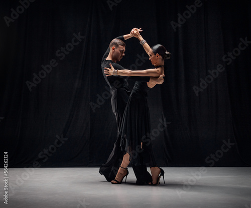 Deep attentive look. Man and woman, professional tango dancers performing in black stage costumes over black background. Concept of hobby, lifestyle, action, motion, art, dance aesthetics