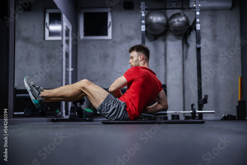 Strengthening the body core and abdominal muscles. A sporty young guy does sit-ups on the side in a sporty red shirt and gray shorts on the floor of a modern gym. Sports routine, healthy living