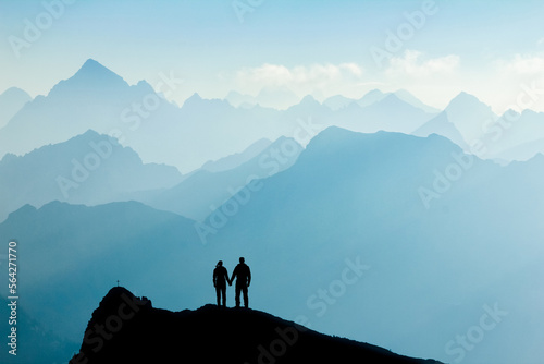 Silhouette Couple of man and woman reaching mountain top enjoying freedom and looking towards blue mountain silhouettes and sunrise. Alps, Allgaeu, Bavaria, Germany. photo