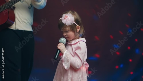 Little girl on stage in vintage dress, she sings into microphone and dances, her father plays acoustic guitar. Color music is shining. Performance on stage. Early development of children