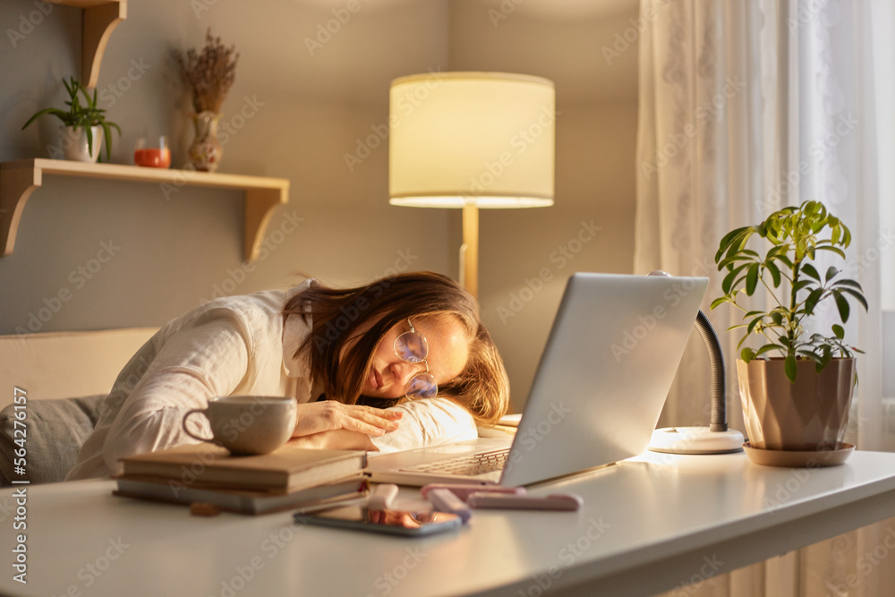 Portrait of tired sleepy Caucasian woman with brown hair sitting at table working on laptop in evening at home, being overworked, sleeping, leaning on her desk. Portrait of tired sleepy Caucasian woman with brown hair sitting at table working on laptop in evening at home, being overworked, sleeping, leaning on her desk.