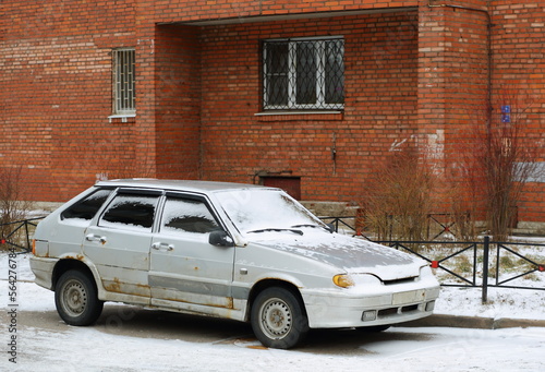 An old rusty silver car covered with snow is parked near a red brick wall, Iskrovsky Prospekt, St. Petersburg, Russia, January 2023