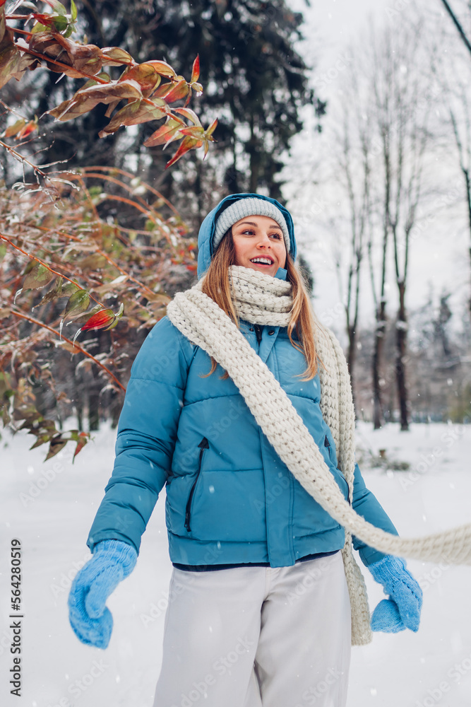 Obraz premium Happy young woman walking in snowy winter park wearing blue coat. Girl enjoys landscape wearing hat and mittens