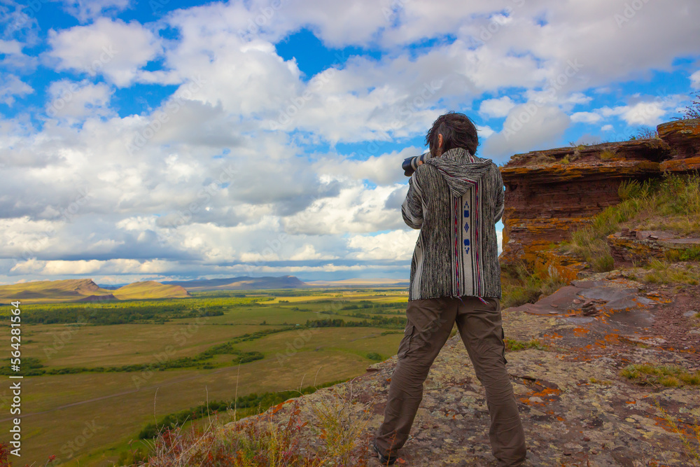 Fototapeta premium A brunet man traveler with a stands on the edge of a cliff and photographs an autumn steppe landscape under cloudy sky. Mountain range Sunduki in Khakassia, Russia. View from the back