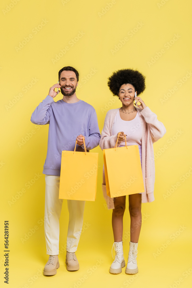 full length of happy interracial couple holding shopping bags and talking on smartphones on yellow background. full length of happy interracial couple holding shopping bags and talking on smartphones on yellow background.