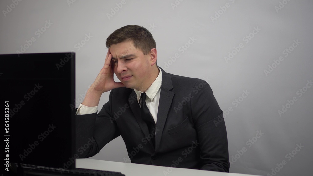 A businessman looks tiredly at a computer monitor and feels hopeless. A young man in a suit works in the office. A businessman looks tiredly at a computer monitor and feels hopeless. A young man in a suit works in the office.