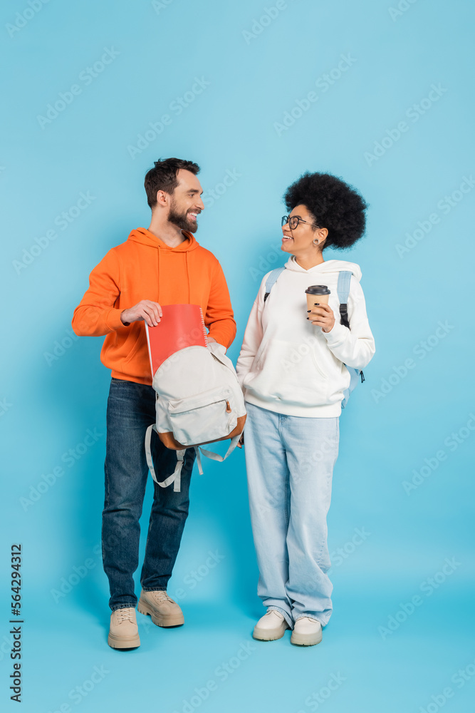 full length of african american woman with takeaway drink looking at bearded boyfriend with backpack and notebook on blue. full length of african american woman with takeaway drink looking at bearded boyfriend with backpack and notebook on blue.