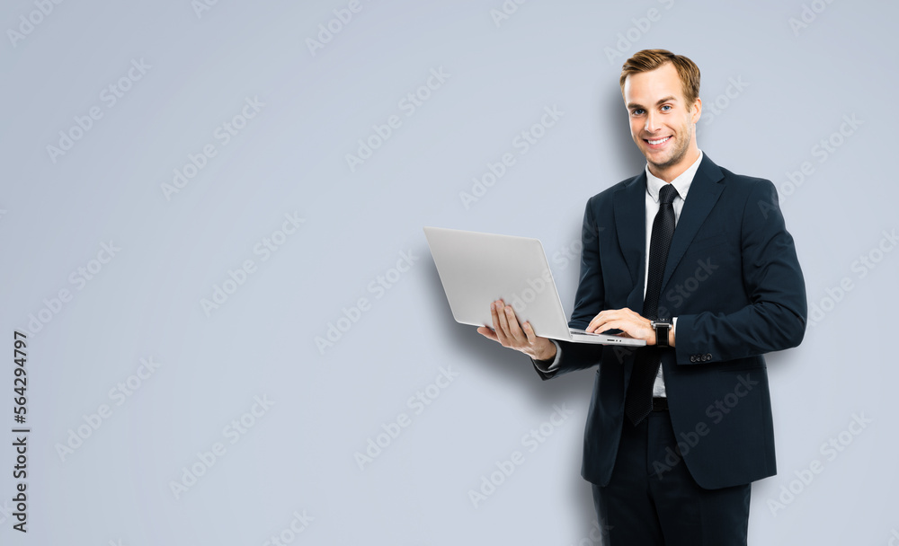 Happy smiling businessman in black confident suit, working with laptop, against grey color background. Handsome young man at studio concept picture. Copy space for ad text. Happy smiling businessman in black confident suit, working with laptop, against grey color background. Handsome young man at studio concept picture. Copy space for ad text.