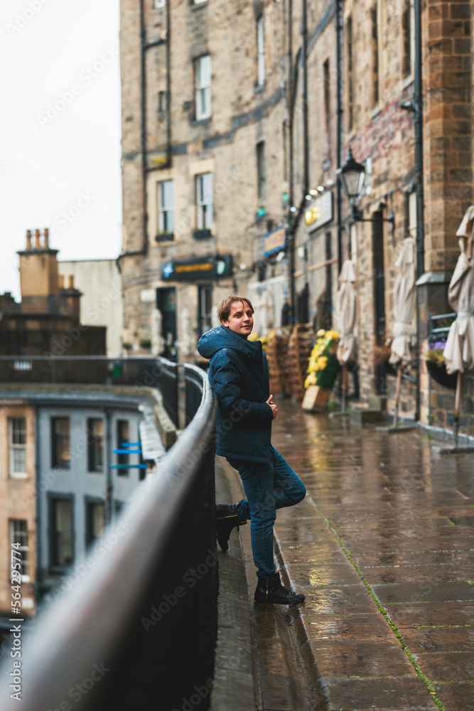 Victoria Street in Edinburgh, the rain drenching his coat as he gazes down the bustling street. Despite the gloomy weather, the city's charm and history are evident in the Victorian architecture