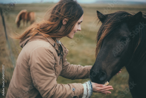 Young woman on the nature in cold day. Tourist female strokes the black beautiful horse in mountains in Iceland. © vadim_key