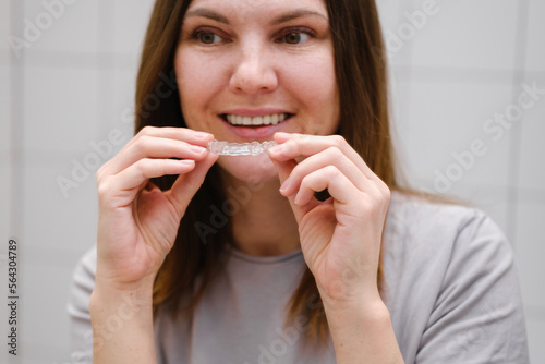 Close up of a smiling woman in front of a mirror in the bathroom holding invisible plastic teeth aligners in hands. Putting on braces. Beautiful and healthy smile. Selective focus.