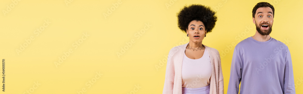 amazed multiethnic couple looking at camera while standing isolated on yellow, banner. amazed multiethnic couple looking at camera while standing isolated on yellow, banner.