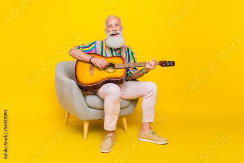 Portrait of optimistic old man with long beard wear colorful shirt sit in armchair playing guitar isolated on yellow color background