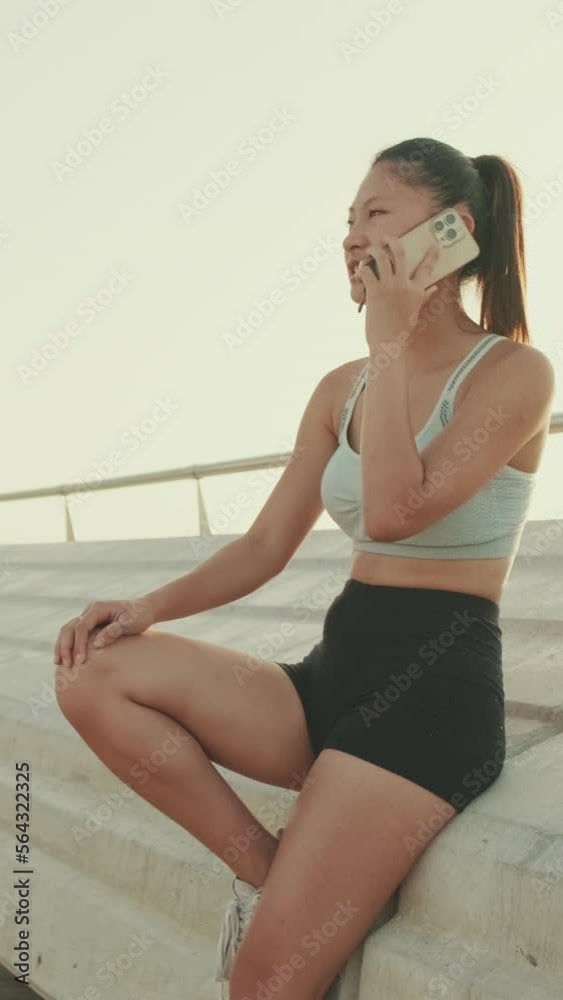 VERTICAL VIDEO: Asian girl in sports top sits on the embankment, talking on cellphone VERTICAL VIDEO: Asian girl in sports top sits on the embankment, talking on cellphone