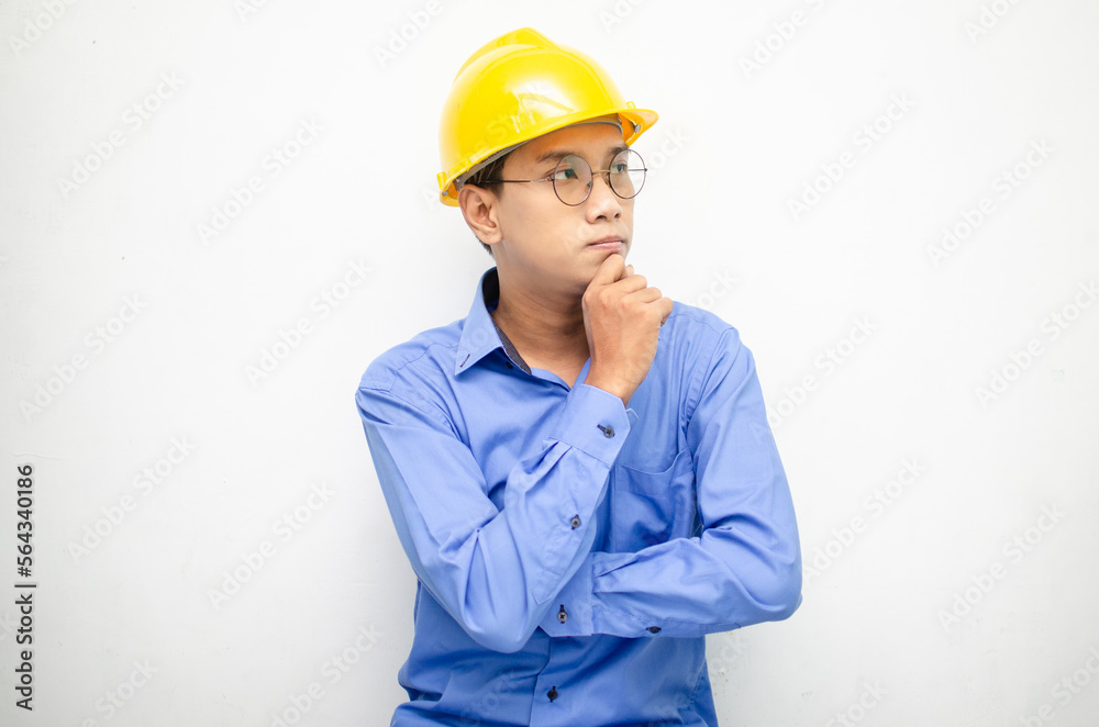 Asian construction worker in blue shirt and yellow safety helmet is seriously thinking with a wondering and thinking gesture and expression.