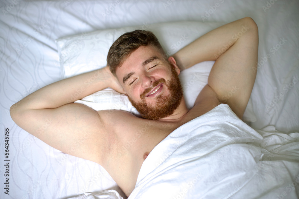 Happy young handsome man is waking up in the morning in bed in bedroom, stretching his arms, body, smiling. Happy young handsome man is waking up in the morning in bed in bedroom, stretching his arms, body, smiling.