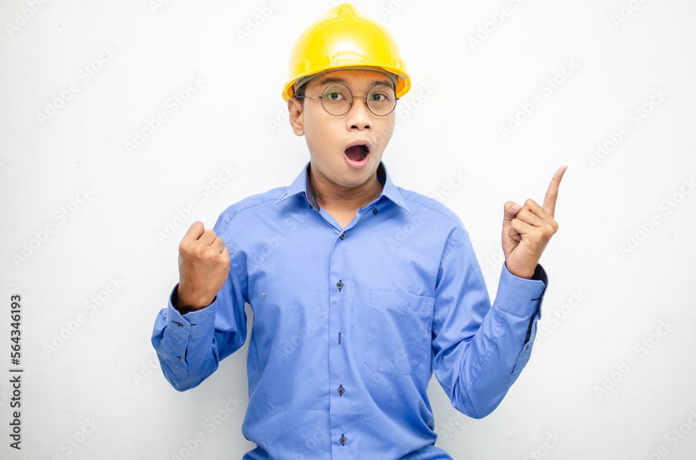 A happy, shocked asian consrtuction worker in blue shirt and yellow safety helmet pointing finger and presenting something ads. 