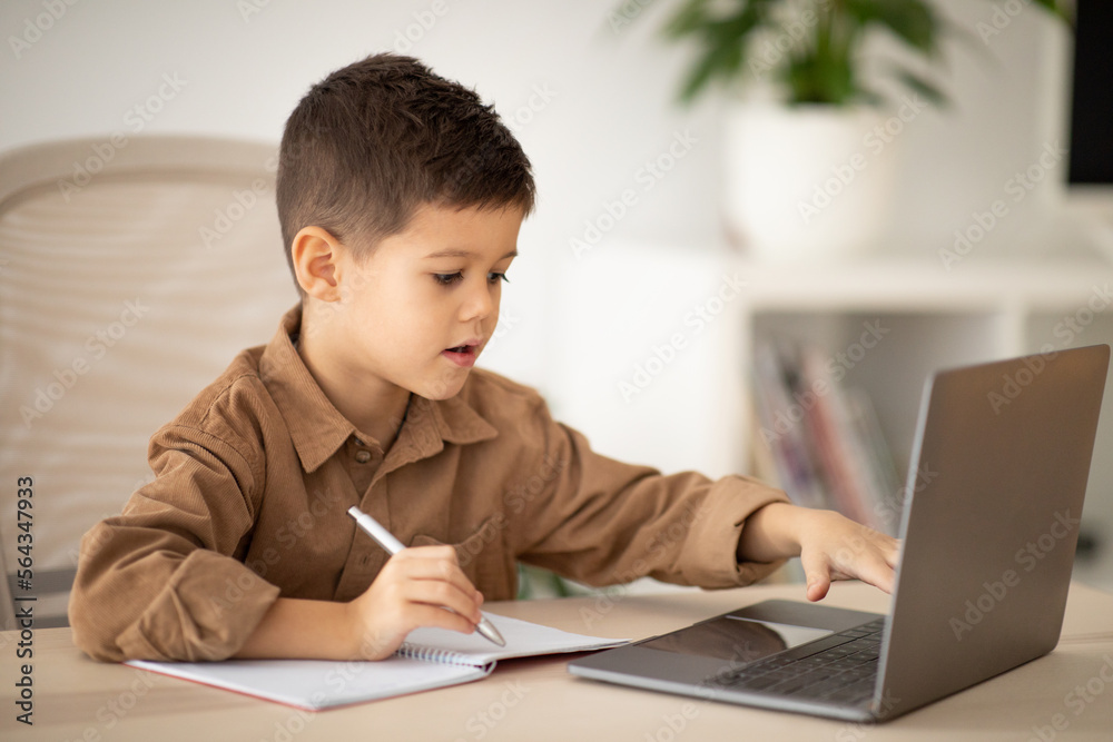 Serious busy little boy sits at table, studies, has video call on laptop, makes notes in room interior Serious busy little boy sits at table, studies, has video call on laptop, makes notes in room interior