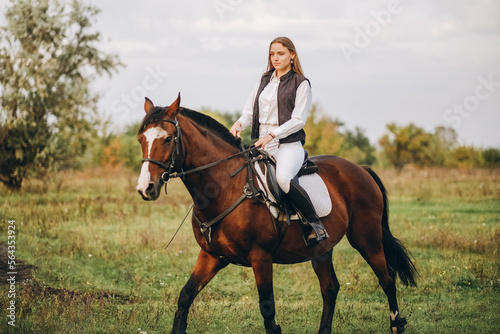 Young beautiful blonde woman jockey rides a brown horse in a meadow at sunset in summer. Preparing for an equestrian competition. © sergo321