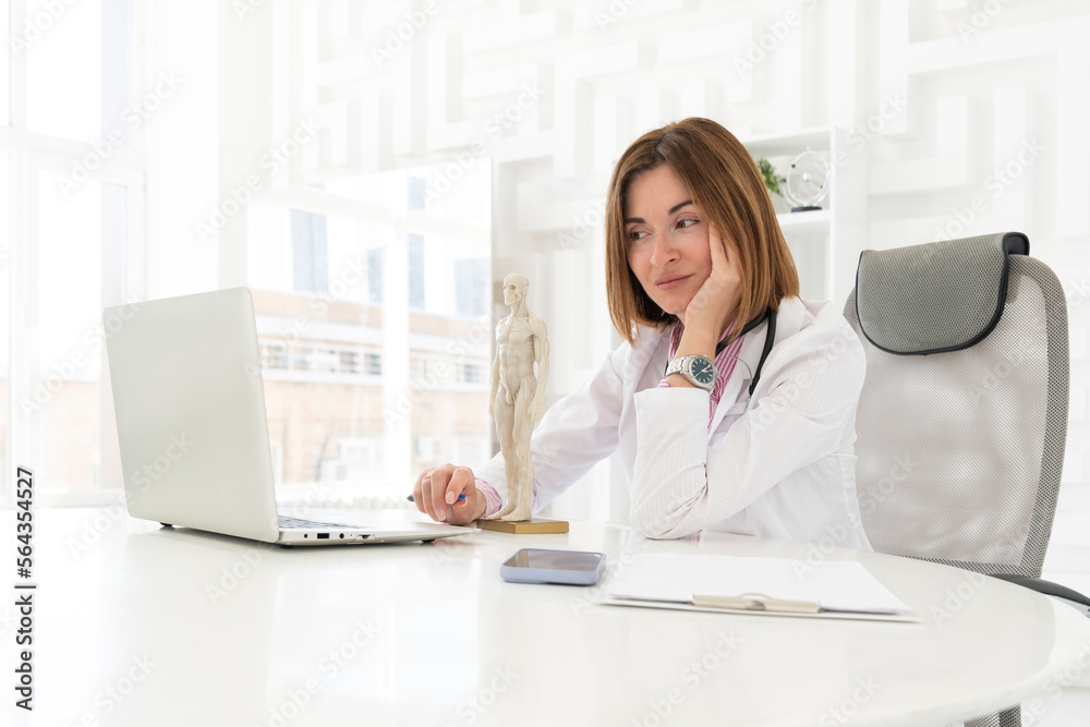 Portrait photo of young and beautiful smiling female doctor in white medical gown sitting and working on laptop in the office of the modern clinic. Online consultation and distant cure concept Portrait photo of young and beautiful smiling female doctor in white medical gown sitting and working on laptop in the office of the modern clinic. Online consultation and distant cure concept