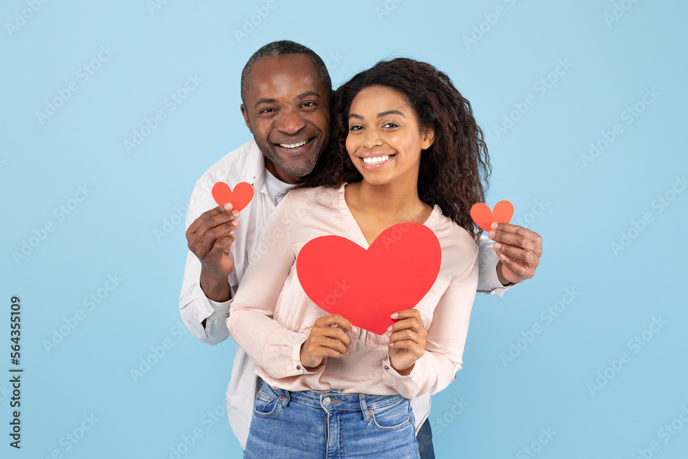 Love is in the air. Portrait of romantic black couple with red paper hearts in hands posing over blue background