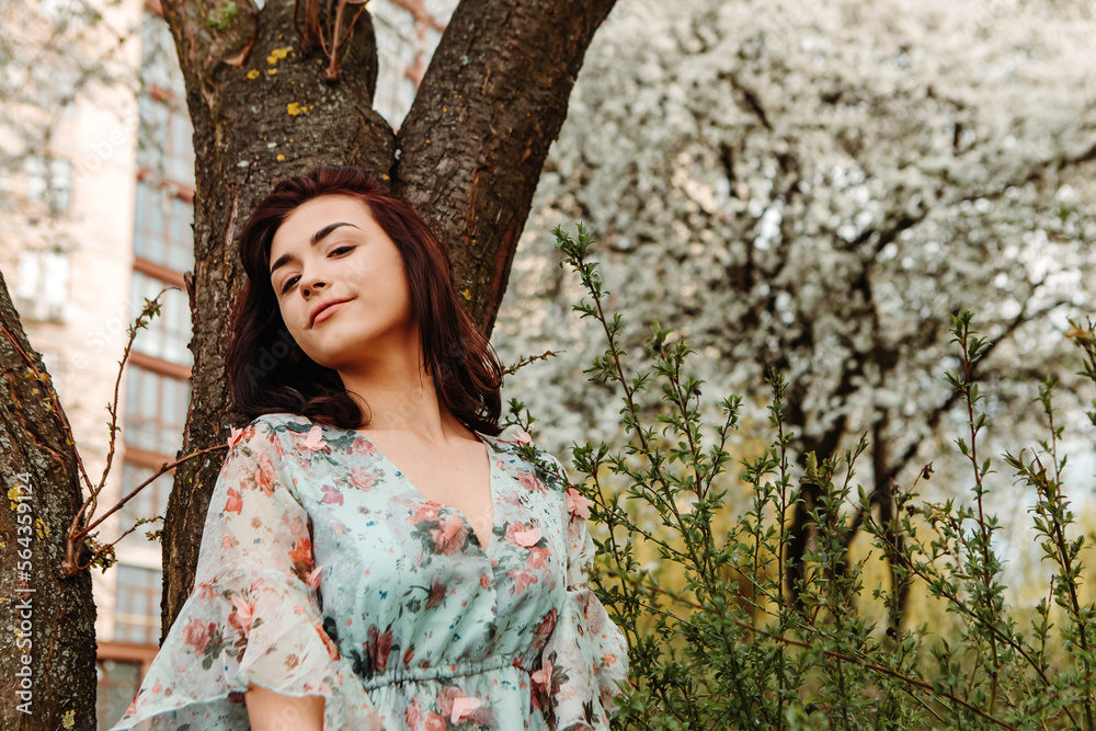 Portrait of charming pretty woman dressed flowery dress posing near apple cherry tree blossoms blooming flowers in the garden park in early spring nature. Fashion, girl model with black hair

