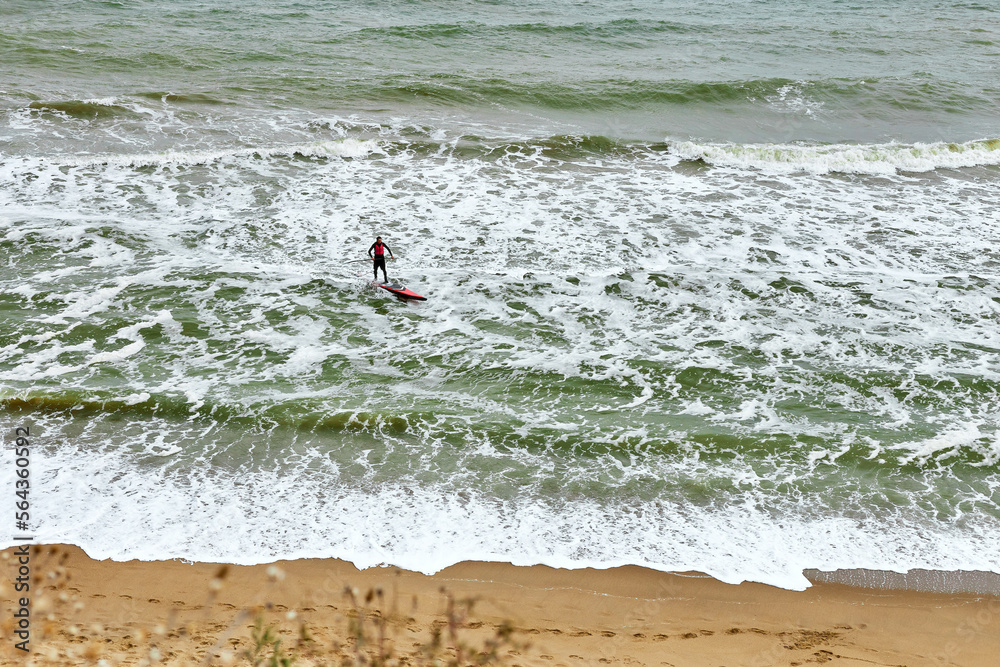 Young brave athlete man surfing by wave on stand up paddle board and catching waves at winter period in sea. Splashes of water. Sea waves, landscape, water sports. View from above.