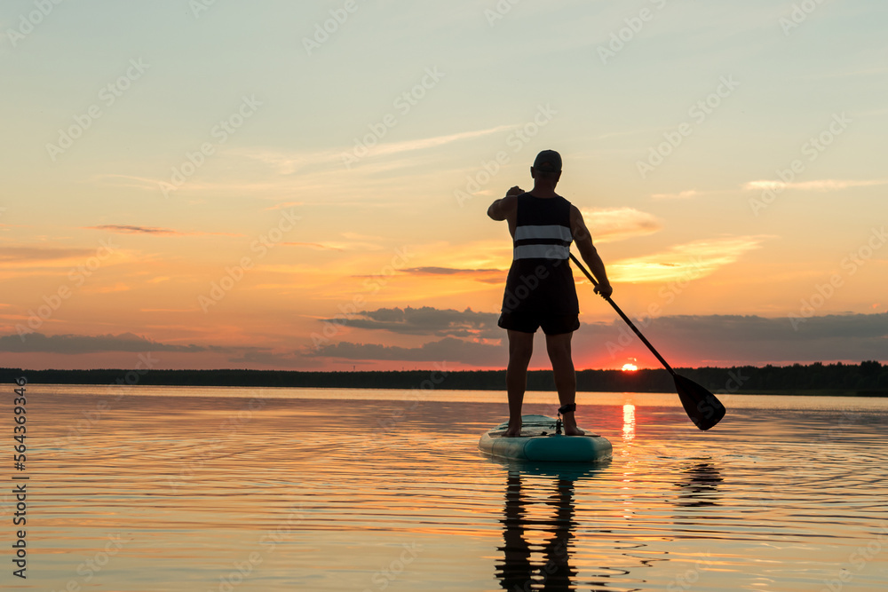 A man in shorts on a sapboard with an oar against the backdrop of a sunset sky swims in the lake in the evening.
