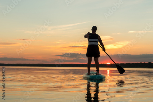 A man in shorts on a sapboard with an oar against the backdrop of a sunset sky swims in the lake in the evening. © finist_4