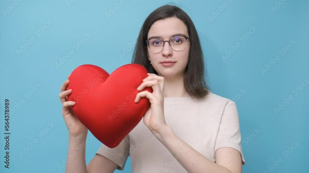 Video portrait of a beautiful student girl in glasses holding a red heart-shaped pillow on a blue background, side view. The pulse of the heart. Love, relationships, Valentine's Day. Video portrait of a beautiful student girl in glasses holding a red heart-shaped pillow on a blue background, side view. The pulse of the heart. Love, relationships, Valentine's Day.