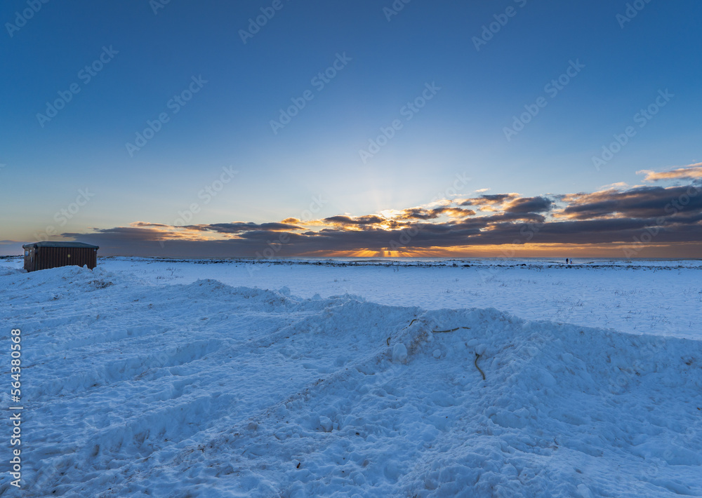 Landscape of the beach area Black sand beach completely covered with snow with golden light of dawn under the clouds and two men walking on the horizon.