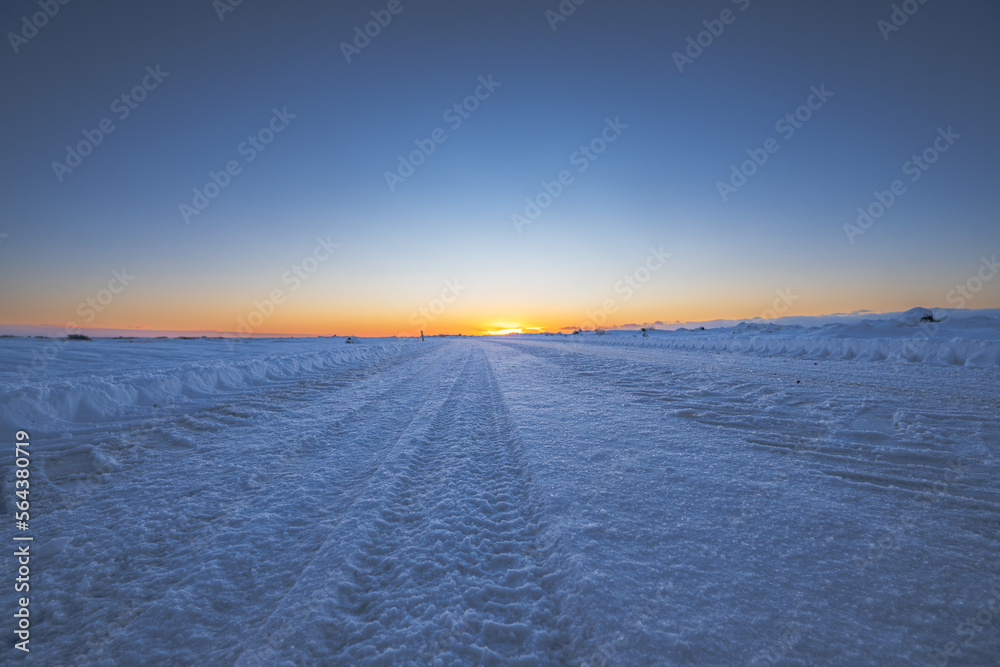 Road and flat snowy landscape with tire tracks going to the horizon with the sun rising in a golden magical Icelandic sunrise.