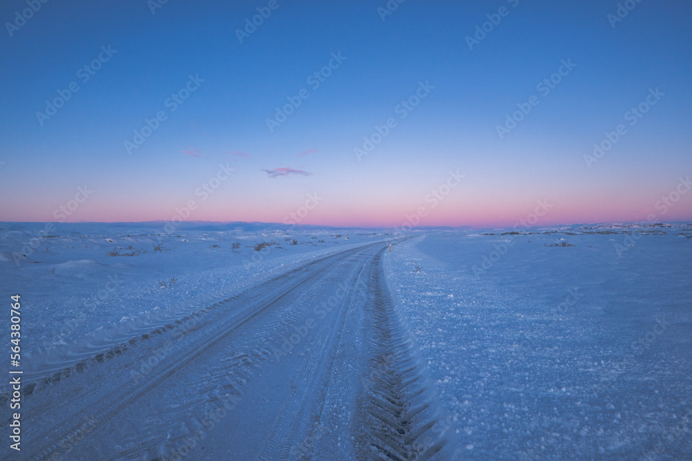 Road and flat snowy landscape with tire tracks going to the horizon with purple magical icelandic sunrise.