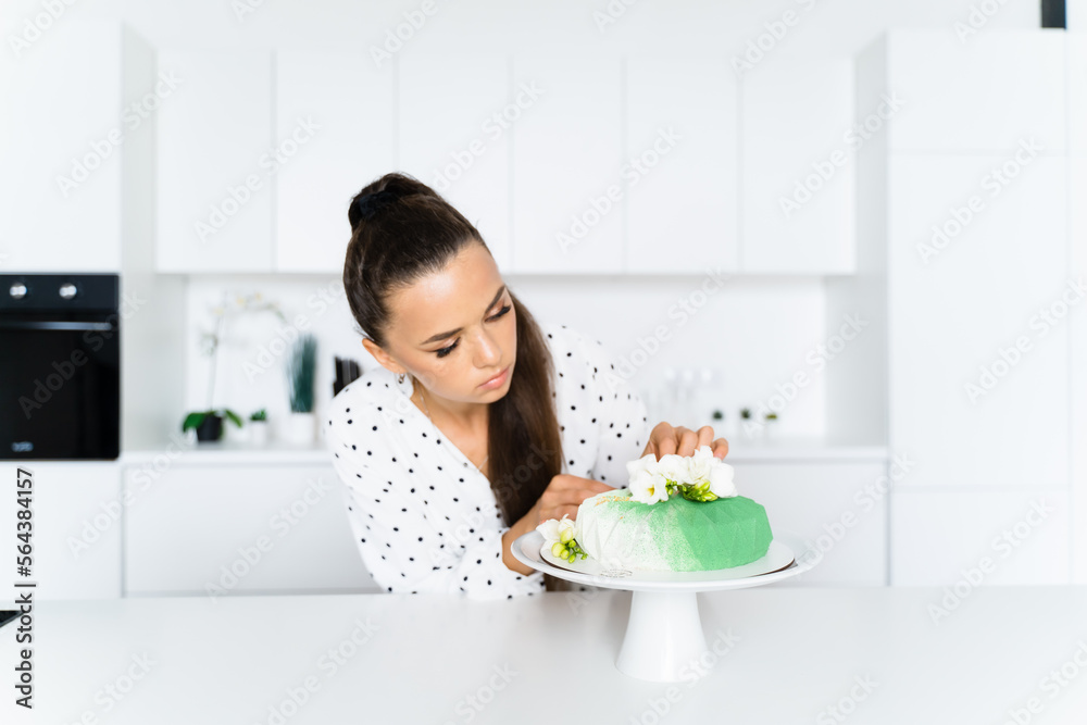 In a white kitchen, a European girl confectioner adjusts a flower on a green mousse cake. Confectioner In a white kitchen, a European girl confectioner adjusts a flower on a green mousse cake. Confectioner
