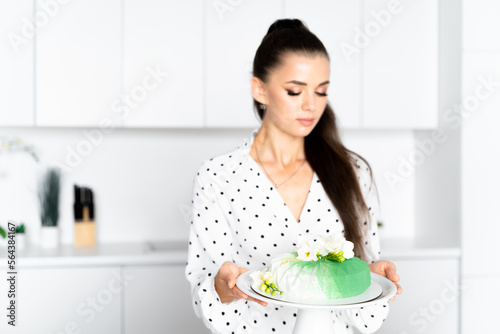 a beautiful girl in a white dress stands in a white kitchen and holds a mousse cake decorated with fresh flowers