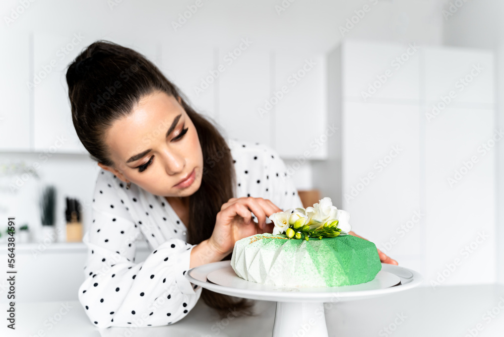 a beautiful girl pastry chef dressed in a white dress with polka dots in a white kitchen decorates a green mousse cake with a white flower a beautiful girl pastry chef dressed in a white dress with polka dots in a white kitchen decorates a green mousse cake with a white flower