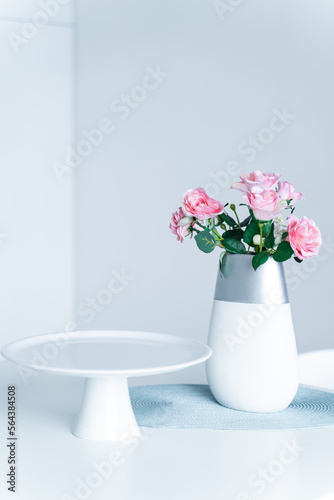 In the white kitchen on the table is an empty white tray for sweets and pink flowers in a vase - roses. Concept, interior, style, fashion, life, romance