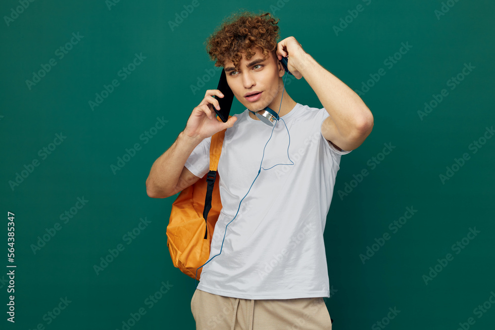 horizontal portrait of a handsome man with curly hair standing on a green background in a gray T-shirt, holding headphones around his neck, talking enthusiastically on the phone horizontal portrait of a handsome man with curly hair standing on a green background in a gray T-shirt, holding headphones around his neck, talking enthusiastically on the phone