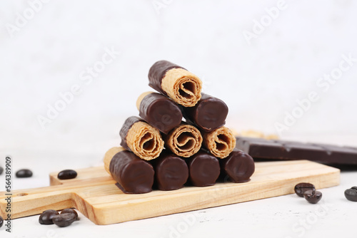 Cutting board with heap of delicious wafer rolls, coffee beans and chocolate on light wooden background, closeup