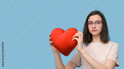 Portrait of a beautiful smiling student girl with glasses holding a bright red heart-shaped pillow
