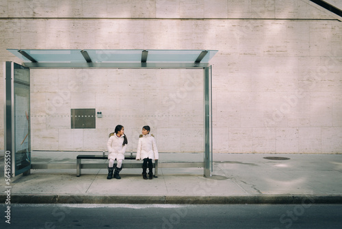 Photography Two asian girls dressed in winter clothe sitting at a bus stop in the city